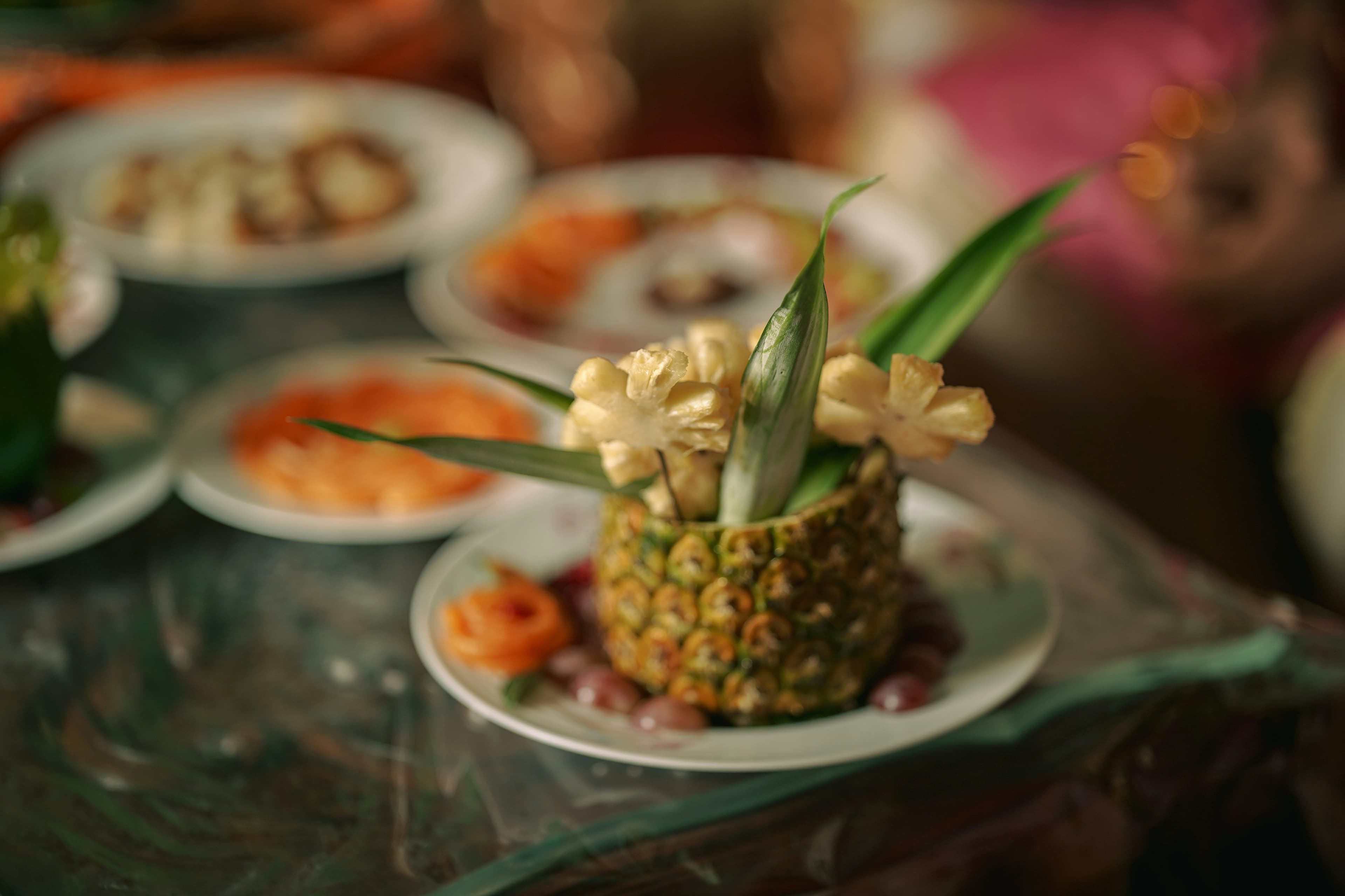 A carved pineapple table centerpiece with pandanus leaves, surrounded by plates of food in soft warm light
