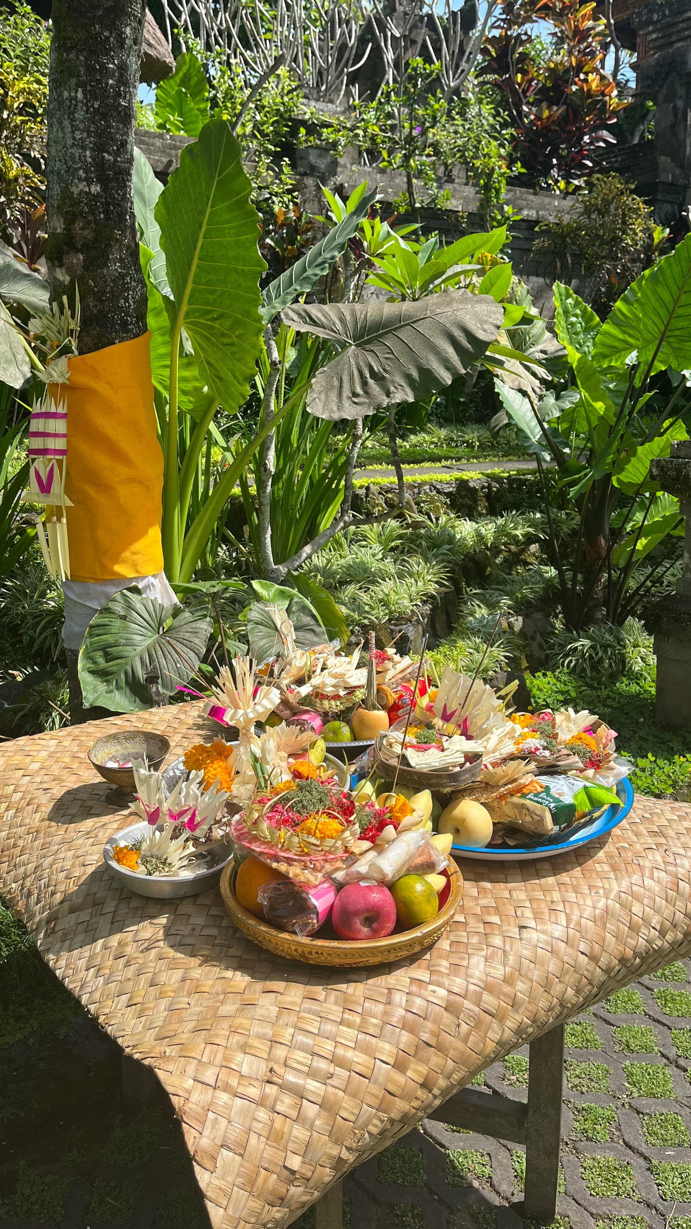 Balinese-style fruit and flower offerings on a woven raffia table, framed by large kalo (taro) leaves in a tropical garden