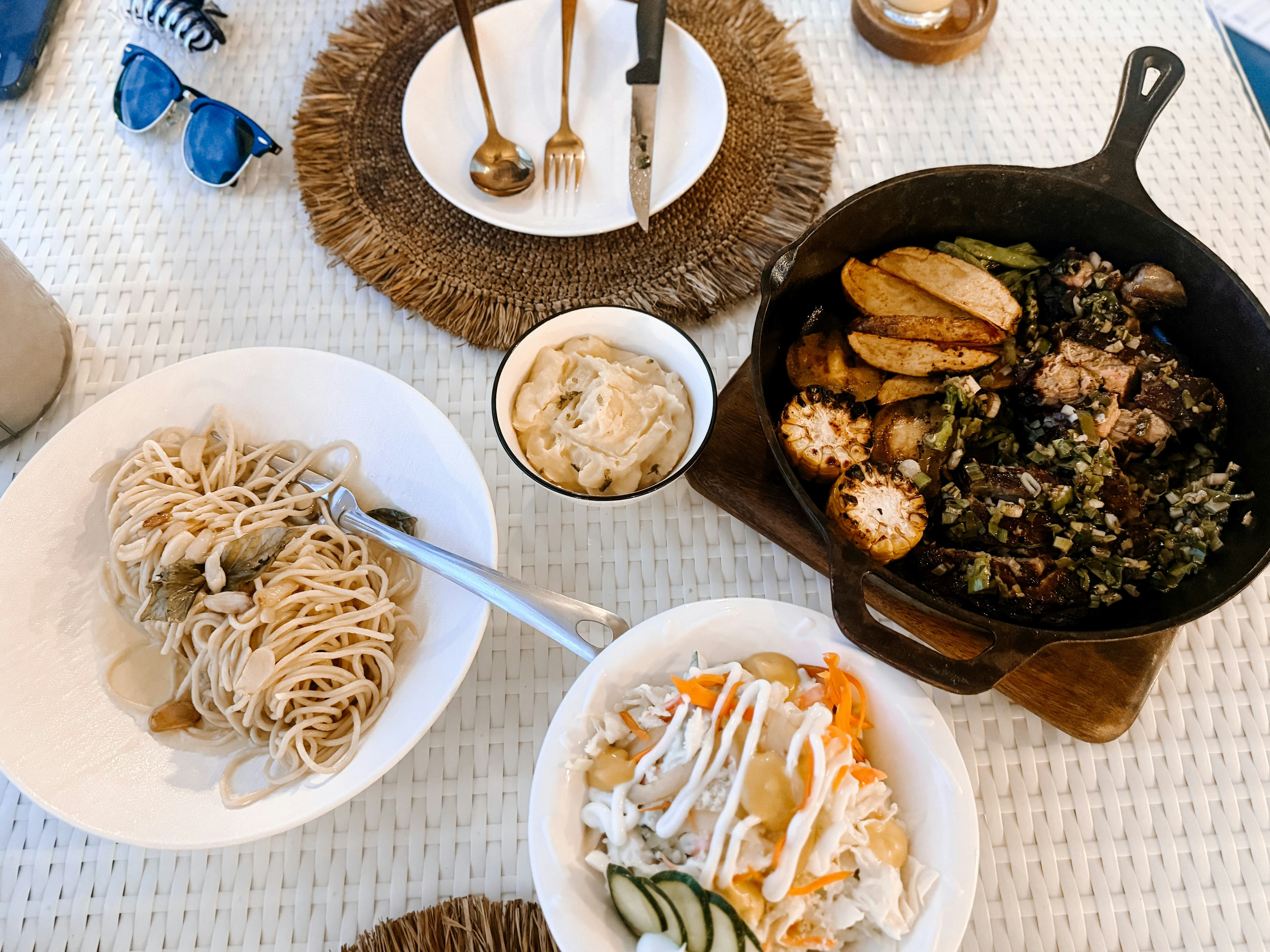 Overhead Polynesian table spread on a woven raffia tablecloth — cast-iron skillet with seasoned greens and protein, rice noodles, slaw, and dipping sauce